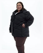 Black Geneva women's plus size mid length synthetic down jacket, three quarter view, hand on hip